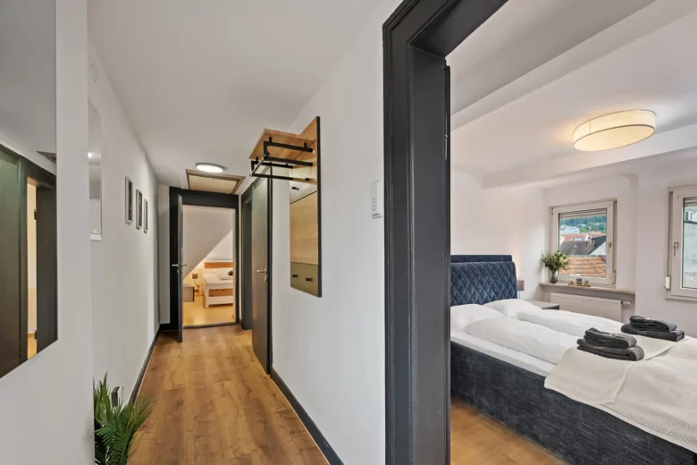 Ein moderner Flur mit Holzboden in dieser Bad Kissinger Ferienwohnung in der Bachstraße führt zu einem hellen Schlafzimmer mit einem großen Bett, zwei Fenstern und frischen Handtüchern. Eine Pflanze schmückt den Flur, und ein weiteres Zimmer ist am Ende des Flurs zu sehen.