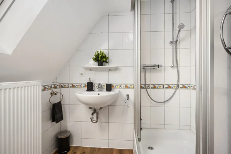 Das kleine geflieste Badezimmer in der Ferienwohnung Bachstraße verfügt über ein Eckwaschbecken, ein Wandregal mit einer Topfpflanze, eine Dusche mit Glastür, einen Handtuchring und ein schwarzes Handtuch sowie einen Heizkörper unter der schrägen Decke.