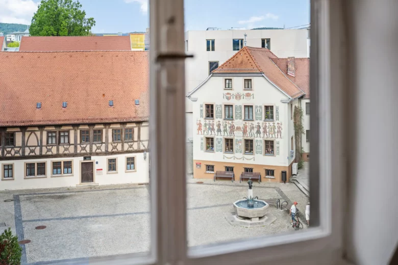 Der Blick aus dem Fenster einer Wohnung in der Bachstraße in Bad Kissingen zeigt Rathausplatz mit einem zentralen Brunnen, einem Fachwerkhaus und einem weißen Gebäude, das mit gemalten Figuren geschmückt ist.