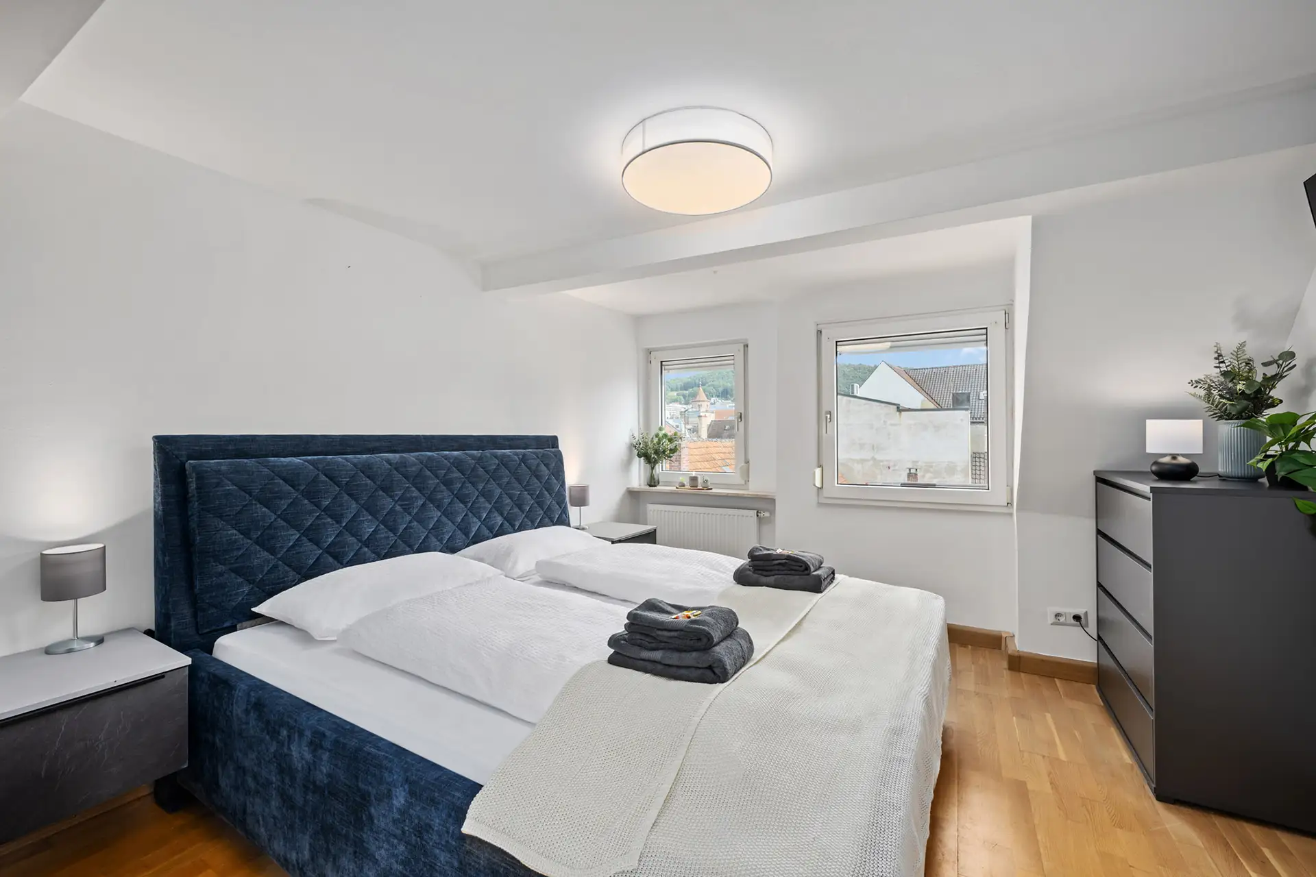 Modernes Schlafzimmer in einer Ferienwohnung in der Bachstraße, Bad Kissingen, mit einem blauen Polsterbett mit weißer Bettwäsche, gefalteten Handtüchern, Nachttischen mit Lampen, einer schwarzen Kommode, einer Pflanze, Holzböden und zwei Fenstern, die Tageslicht hereinlassen.