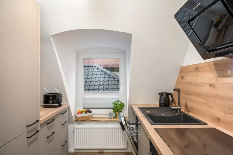 Eine kompakte, moderne Küche in App.3, Kirchgasse, mit hellen Holzarbeitsplatten, beigen Schränken, einer schwarzen Spüle, Geräten, einer Obstschale und einer Basilikum-Topfpflanze an einem Fenster mit Blick auf ein Ziegeldach.