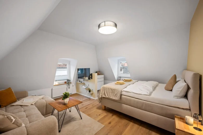 Das gemütliche Schlafzimmer im Dachgeschoss von App.3, Kirchgasse, verfügt über schräge Decken, ein beiges Bett, ein hellbraunes Sofa und einen Couchtisch aus Holz. Zwei kleine Fenster lassen natürliches Licht auf Pflanzen, Bücher, eine Kerze und neutralfarbene Kissen und Decken fallen.