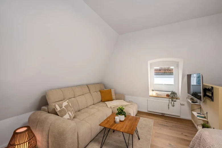 Das gemütliche Wohnzimmer in App.3 in der Kirchgasse ist mit einem beigen Sektionssofa, einem Couchtisch aus Holz, einer Topfpflanze und einem Fernseher an einem kleinen Bogenfenster ausgestattet. Helle Holzböden und neutrales Dekor schaffen eine warme Atmosphäre.