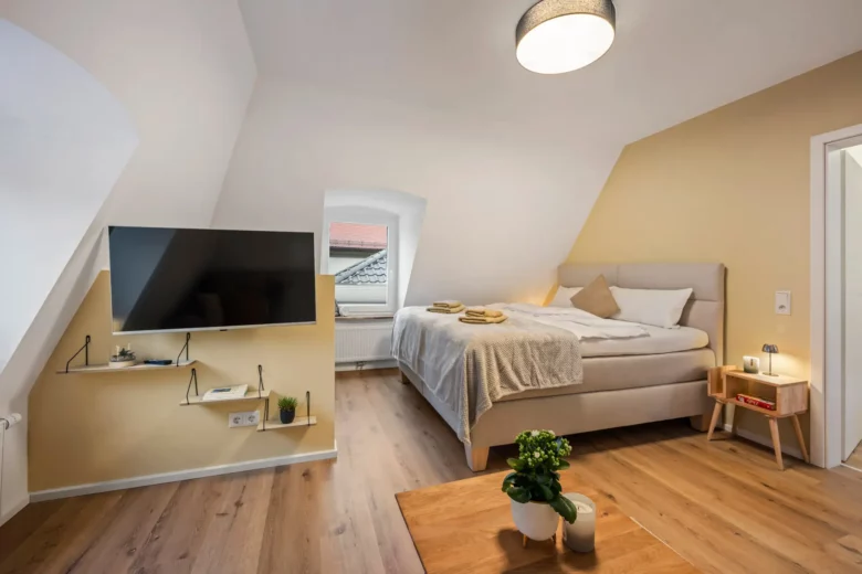 Gemütliches Schlafzimmer in App.3, Kirchgasse, mit Dachschräge, Doppelbett mit beiger Bettwäsche, Wandfernseher, schwebenden Regalen, Nachttisch mit Lampe und einem kleinen Tisch mit Pflanzen auf Holzboden. Natürliches Licht durch ein Rundbogenfenster.