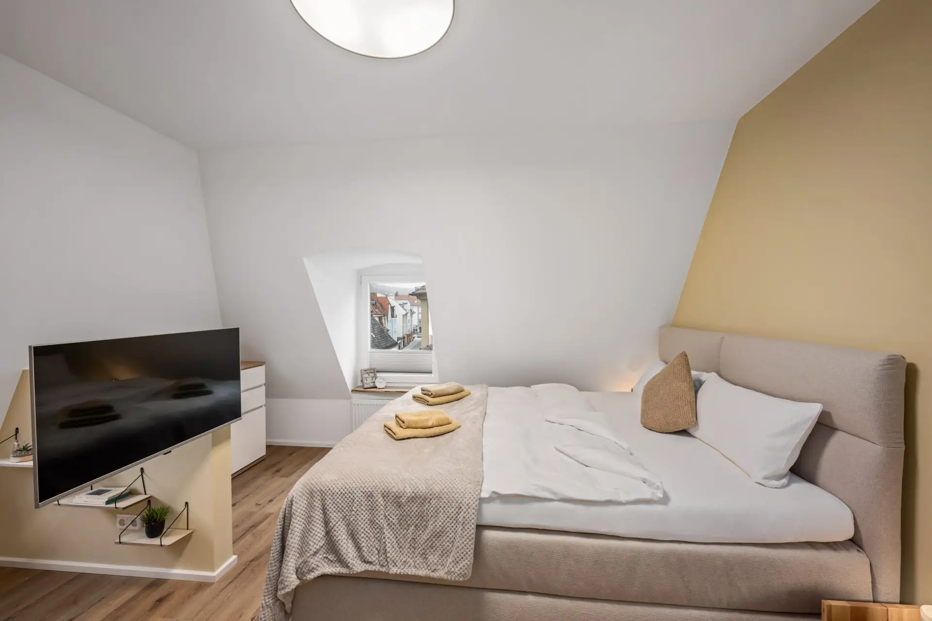 Modernes Schlafzimmer in der Kirchgasse App.3 mit einem großen Bett, beiger Bettwäsche, einer schrägen Decke, einem an der Wand montierten Fernseher und einem kleinen Fenster mit Blick auf die Stadt. Helle Holzböden und eine gelbe Akzentwand vervollständigen den gemütlichen Raum.
