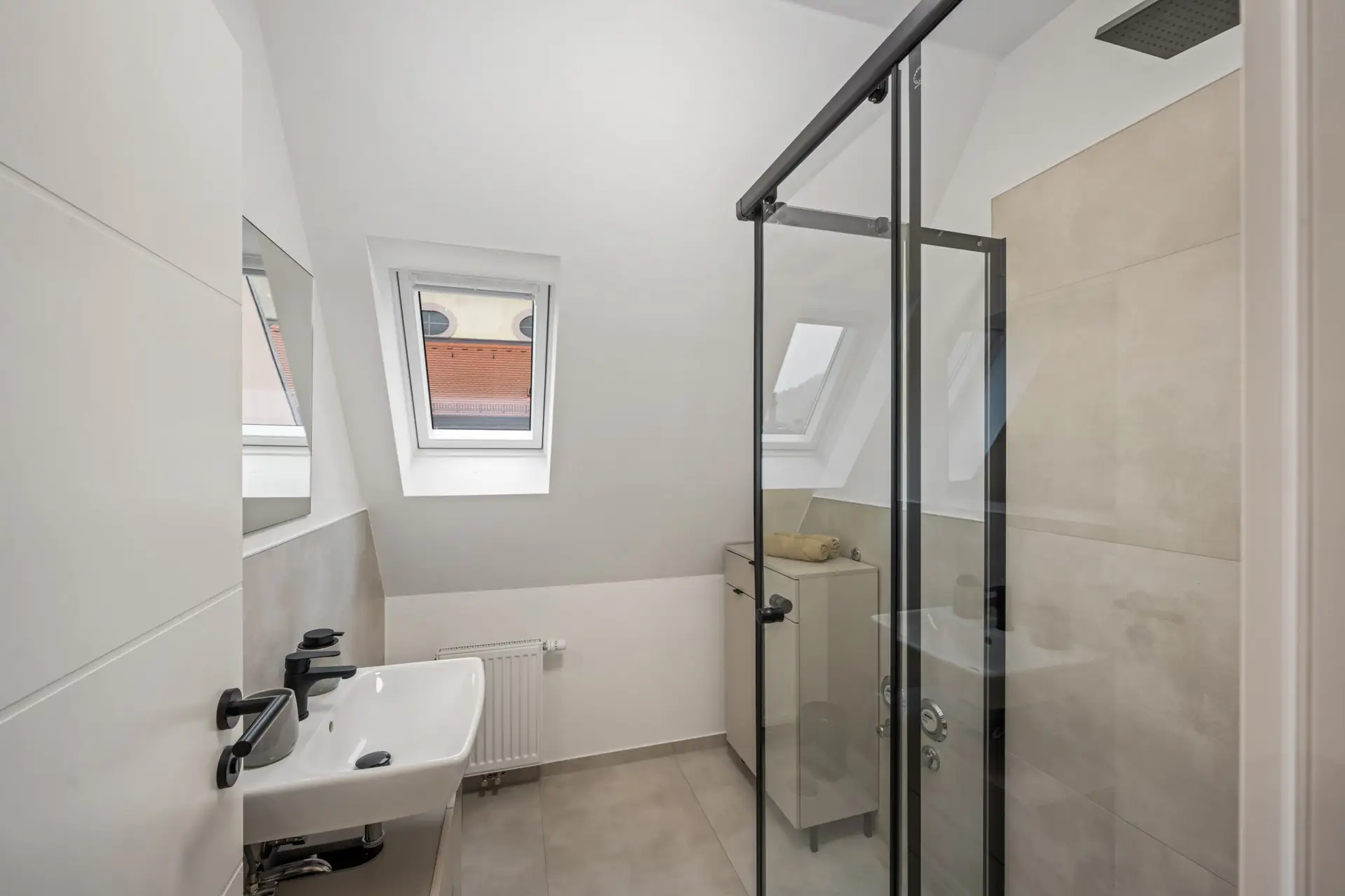 Modernes Badezimmer in der Kirchgasse App.3 mit abgewinkelter Decke und zwei Oberlichtfenstern. Ausgestattet mit einer gläsernen Dusche, einem Wandwaschbecken mit schwarzen Armaturen, beigen Fliesen, einem kleinen Schrank und einem Heizkörper unter den Fenstern.