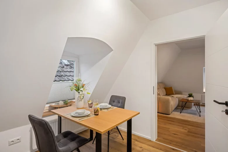 Eine kleine Essecke in App.3 mit einem Holztisch für zwei Personen, grauen Stühlen und einer Vase mit Blumen am Fenster. Ein offener Durchgang führt in ein gemütliches Wohnzimmer mit Sofa und Holzboden in der Kirchgasse.