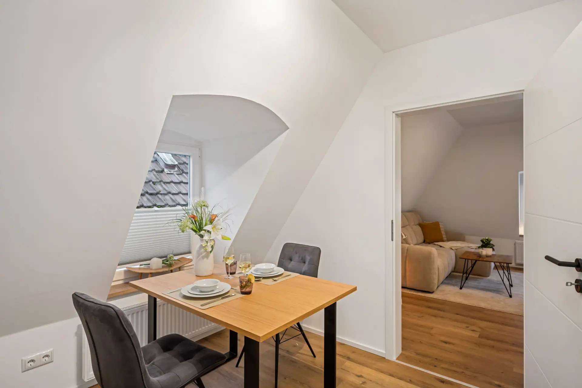 Eine kleine Essecke in App.3 mit einem Holztisch für zwei Personen, grauen Stühlen und einer Vase mit Blumen am Fenster. Ein offener Durchgang führt in ein gemütliches Wohnzimmer mit Sofa und Holzboden in der Kirchgasse.